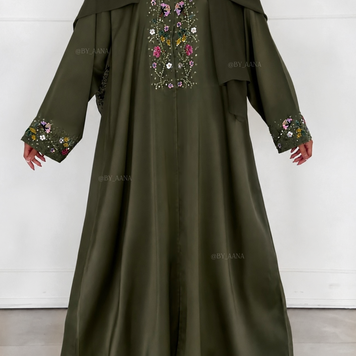 A khaki green abaya dress with a matching headscarf, featuring colorful floral embroidery on the chest and cuffs, shown on a model against a white paneled wall.