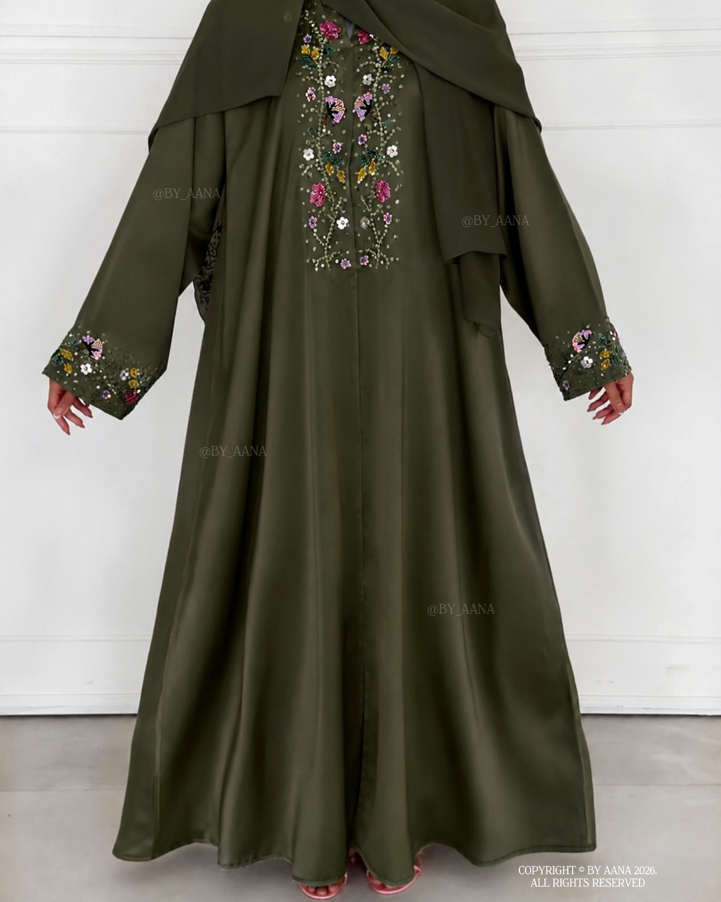 A khaki green abaya dress with a matching headscarf, featuring colorful floral embroidery on the chest and cuffs, shown on a model against a white paneled wall.
