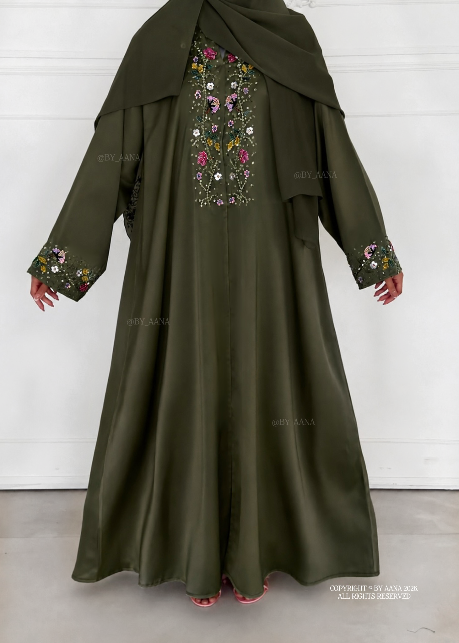 A khaki green abaya dress with a matching headscarf, featuring colorful floral embroidery on the chest and cuffs, shown on a model against a white paneled wall.